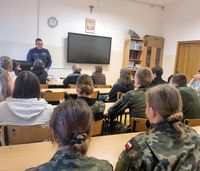 lekcja w klasie o profilu mundurowym, policjant stoi za biurkiem a młodzież siedzi na krzesłach przy ławkach,