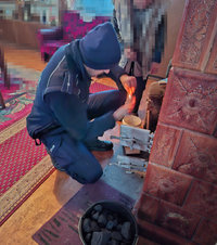 policjant rozpala ogień w piecu kaflowym, za nim stoi starsza kobieta