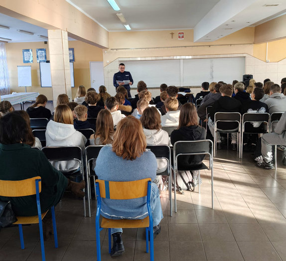 policjant stoi przed młodzieżą siedząca na krzesłach i czyta tekst ustawy