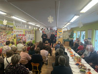 Spotkanie dzielnicowych z babciami i dziadkami przedszkolaków. Dwaj policjanci stoją przed osobami siedzącymi na krzesełkach.