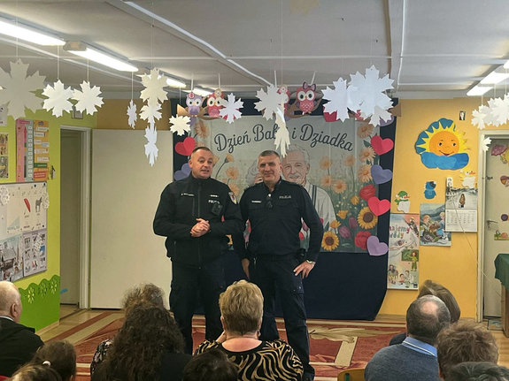 Spotkanie dzielnicowych z babciami i dziadkami przedszkolaków. Dwaj policjanci stoją przed osobami siedzącymi na krzesełkach.