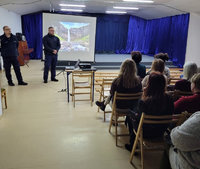 dwaj policjanci stoją przed siedzącymi na krzesłach rodzicami uczniów w czasie spotykania profilaktycznego