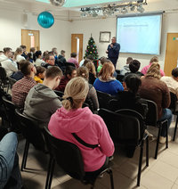 policjant z mikrofonem stoi przed siedzącymi na krzesłach uczniami podczas spotkania profilaktycznego