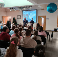 policjant z mikrofonem obok niego policjantka, stoją przed siedzącymi na krzesłach uczniami podczas spotkania profilaktycznego