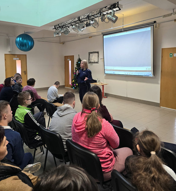 policjantka z mikrofonem stoi przed siedzącymi na krzesłach uczniami podczas spotkania profilaktycznego