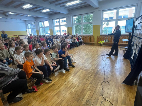 policjantka podczas spotkania profilaktycznego stoi przed dziećmi, które siedzą na ławeczkach