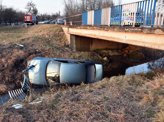 osobowe audi leży na boku w rzece, na moście stoi radiowóz a w tle za mostem inne pojazdy