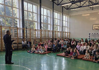 przed siedzącymi na podłodze i krzesełkach uczniami stoi policjant z mikrofonem w czasie spotkania profilaktycznego