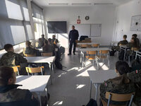 policjant stoi przed siedzącą na krzesłach w ławkach młodzieżą w czasie lekcji