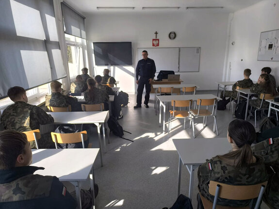 policjant stoi przed siedzącą na krzesłach w ławkach młodzieżą w czasie lekcji