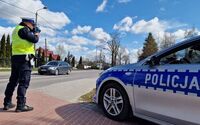 policjant trzyma miernik prędkości, stoi obok radiowozu zaparkowanego na poboczu drogi