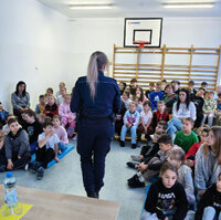 policjanta stoi przed siedzącymi na ławeczkach uczniami