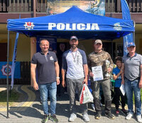 dwuosobowa męska drużyna zawodów wędkarskich w obecności dwóch organizatorów zawodów stoją pod namiotem z napisem Policja, pozują do zdjęcia po wręczeniu nagród za uczestnictwo w zawodach