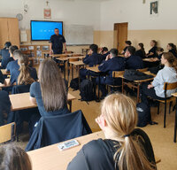 spotkanie profilaktyczne, policjant w szkolnej klasie stoi przy biurku, na przeciwko na krzesłach w ławkach siedzą uczniowie