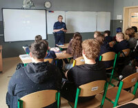 spotkanie profilaktyczne, policjantka w szkolnej klasie stoi przy biurku, na przeciwko na krzesłach w ławkach siedzą uczniowie
