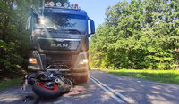 Na drodze leży przewrócony motocykl, którego kierujący uderzył w ciężarówkę. Ciężarówka stoi na drodze za motocyklem.