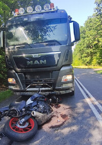 Na drodze leży przewrócony motocykl, którego kierujący uderzył w ciężarówkę. Ciężarówka stoi na drodze za motocyklem.