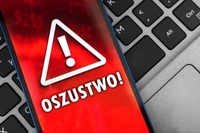 na klawiaturze laptopa leży telefon komórkowy. Na jego wyświetlaczu umieszczony jest trójkątny znak ostrzegawczy oraz napis oszustwo