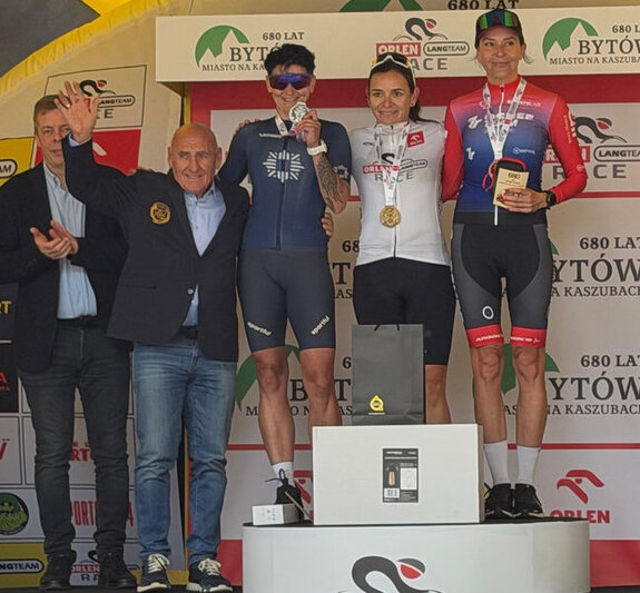 na podium stoją trzy kobiety, zwyciężczynie zawodów kolarskich